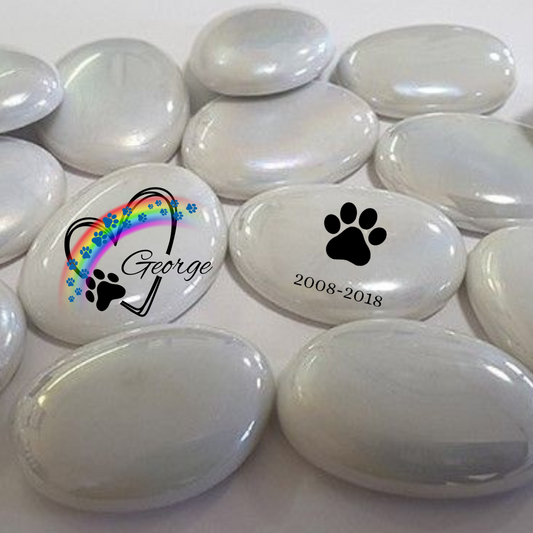 "Conjunto de guijarros de cristal personalizados, mostrando un guijarro con el diseño frontal que incluye el nombre del perro, un arcoíris con azules huellas y un corazón, mientras que otro guijarro muestra el diseño trasero con las fechas personalizadas grabadas."