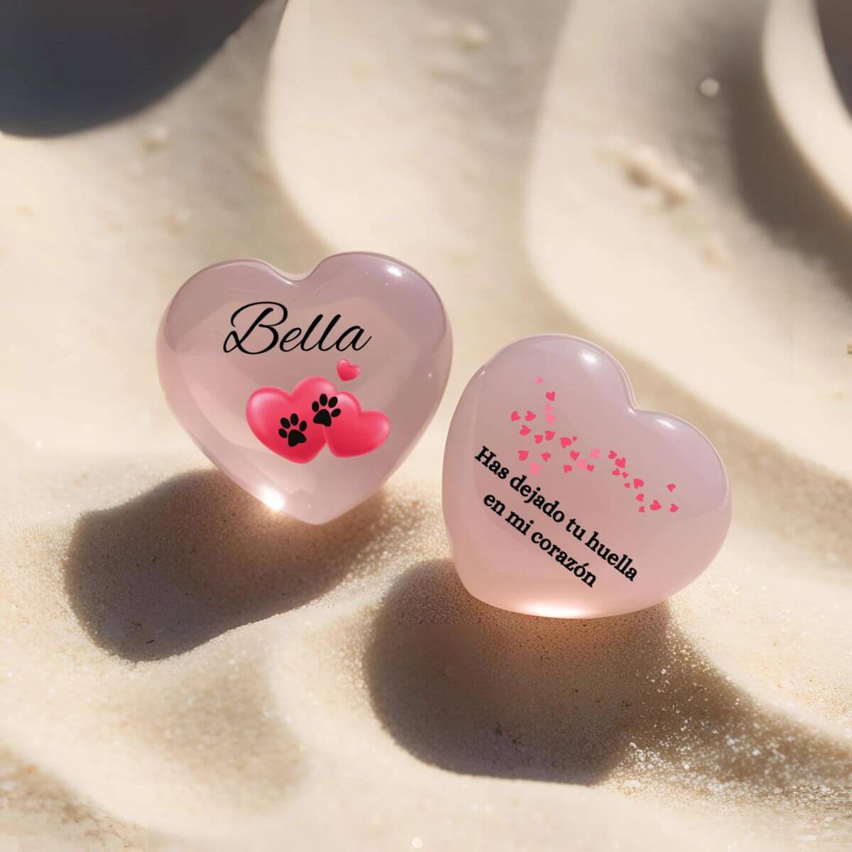 Corazones de cuarzo rosa personalizados con nombre y frase “Has dejado tu huella en mi corazón”, fotografiados sobre arena suave

