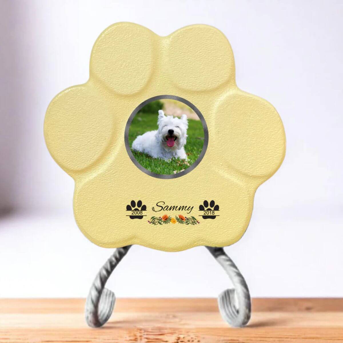 Lápida con forma de huella para mascotas, hecha de piedra amarillo, tamaño pequeño con soporte. Personalizada con foto, nombre y fechas importantes del perro o gato. Ejemplo de homenaje conmemorativo único.

