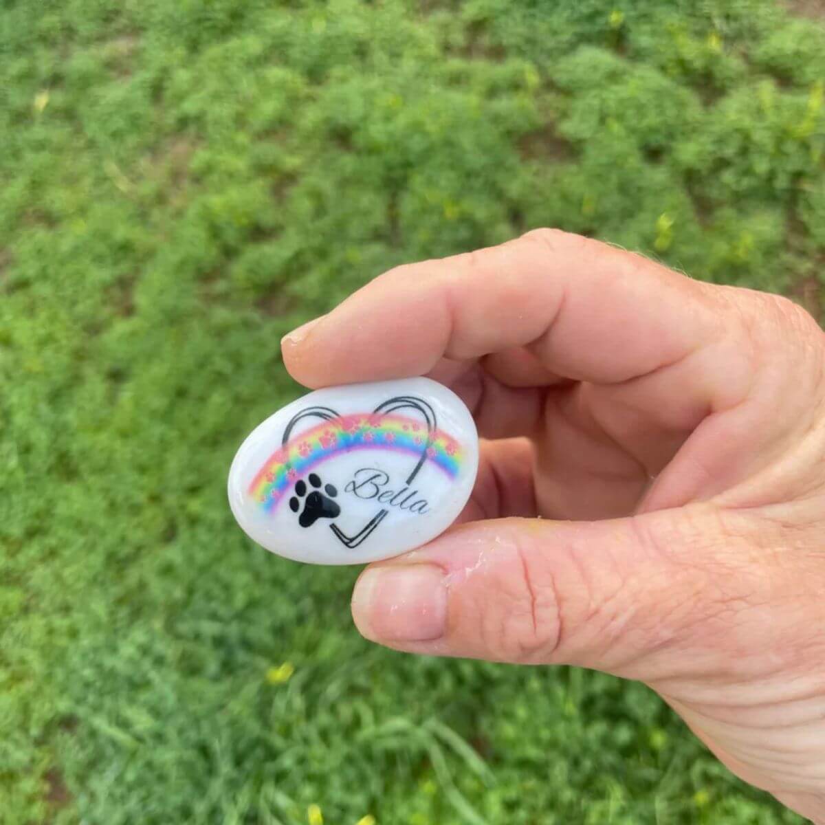 Guijarro de cristal personalizado sostenido entre los dedos, con diseño de huellas rosas, nombre y arcoíris. Regalo conmemorativo ideal para amantes de mascotas.