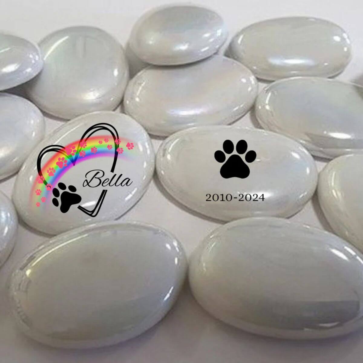 Guijarros de cristal blanco con diseño personalizado: uno con nombre Bella, arcoíris y huellas, y otro con huella negra y fechas. Conmemorativo para mascotas.