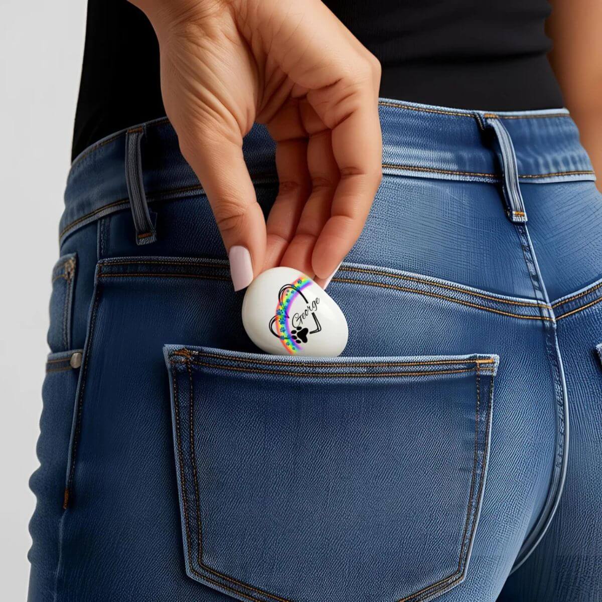 Guijarro de cristal personalizado con nombre George y huellas azules, siendo guardado en el bolsillo trasero del pantalón vaquero. Recuerdo portátil y emotivo para mascotas.
