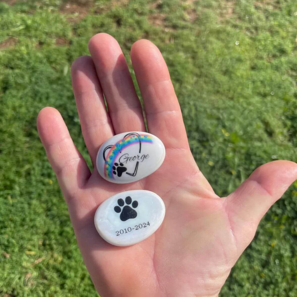 Guijarros de cristal personalizados con huellas azules, uno con nombre “George” y arcoíris, otro con huella negra y fechas. Ambos en la palma de una mano sobre fondo verde.