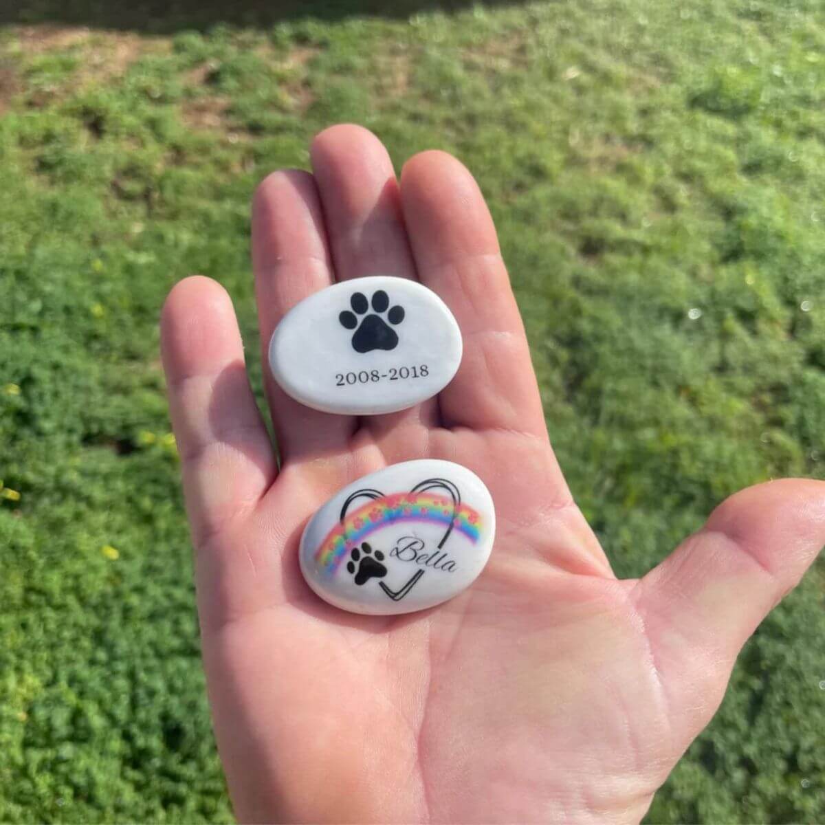 Guijarros de cristal personalizados sostenidos en una mano, uno con nombre y arcoíris y otro con huella y fechas. Recuerdo conmemorativo para mascotas.